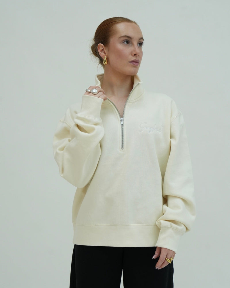 Woman wearing a cream-colored pullover with a zipper against a plain background