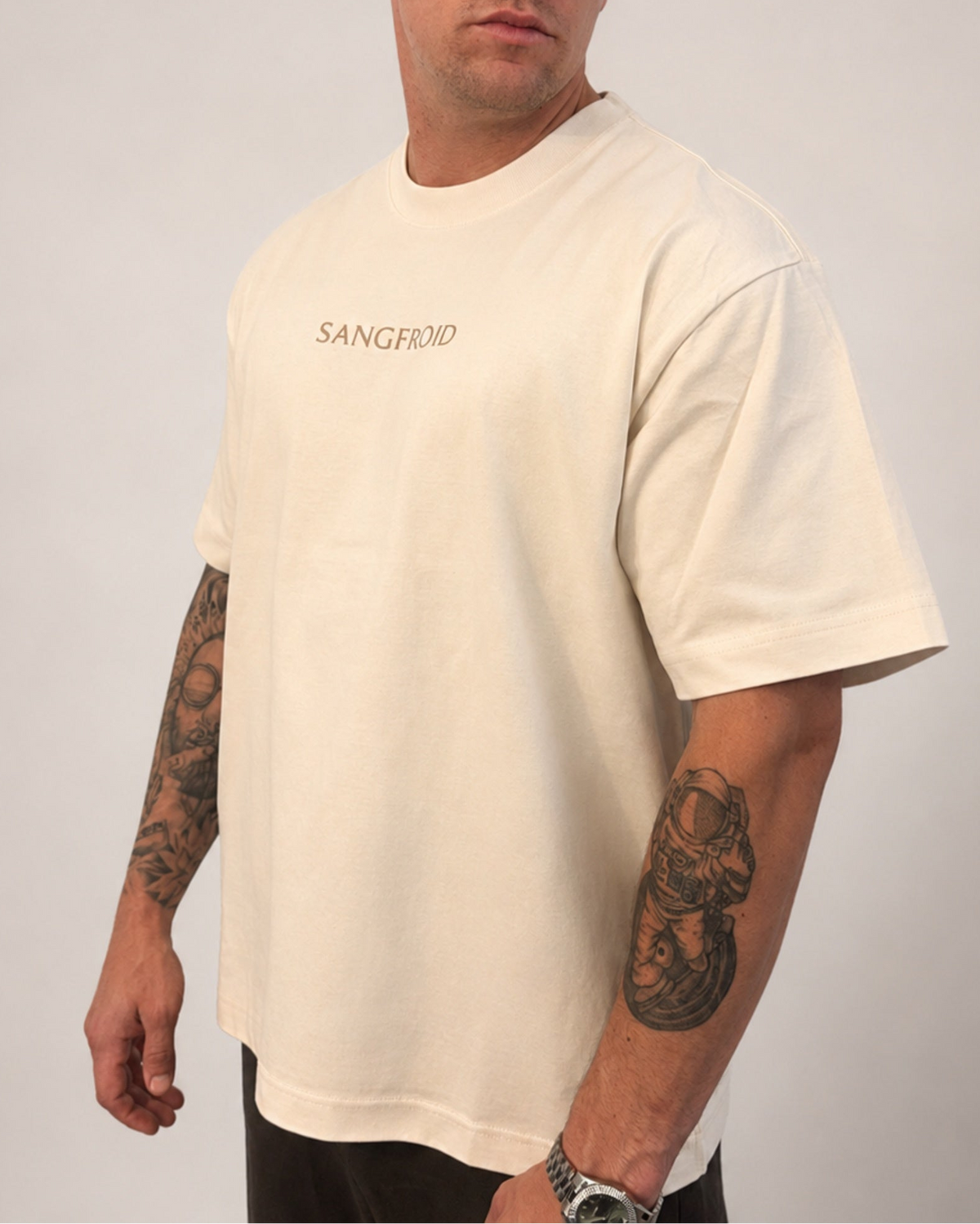 A man wearing an oversized cream-colored t-shirt with 'SANGFROID' embroidered in golden brown on the chest, shown from the neck down against a light gray background.