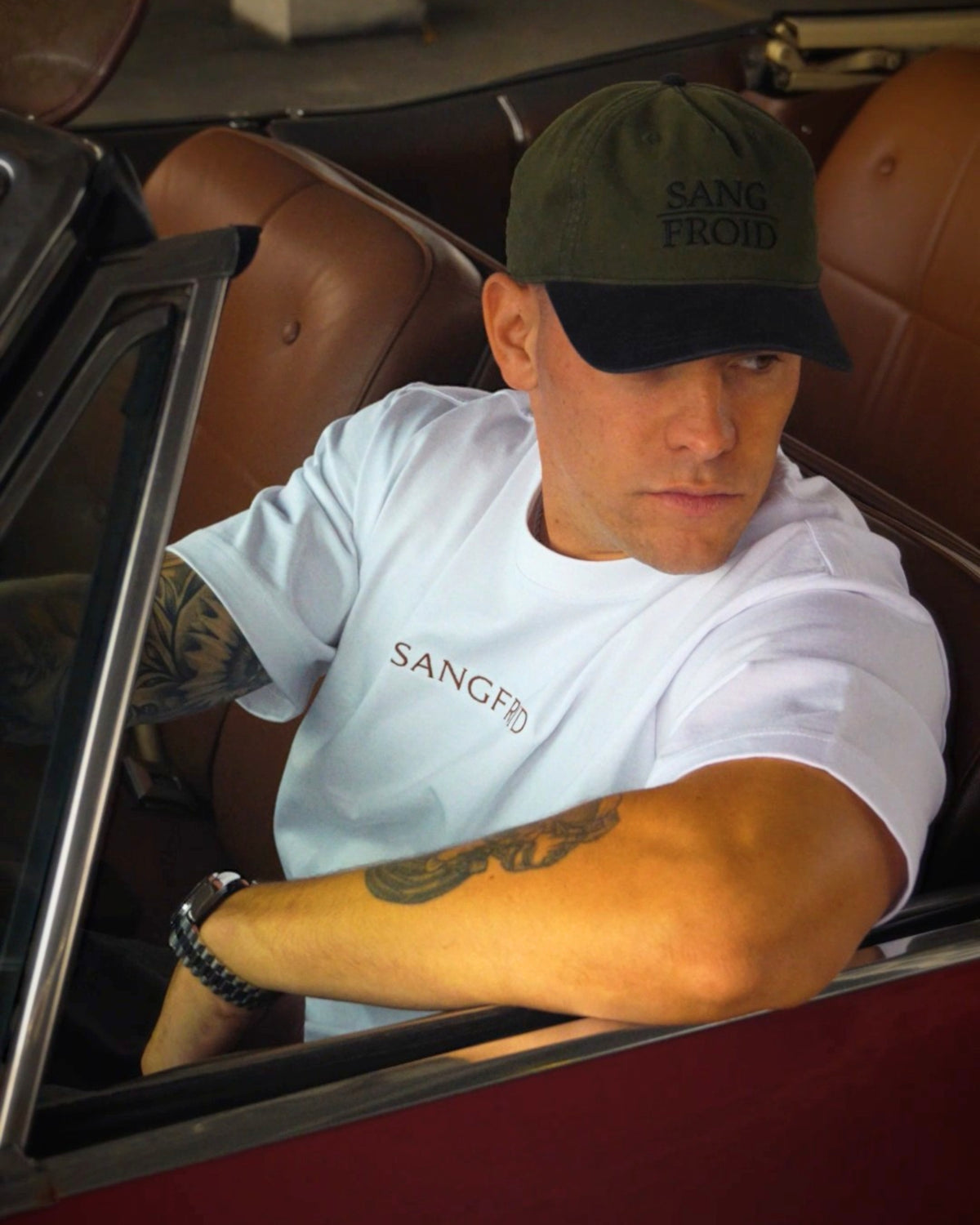 A man sitting in a brown leather convertible car wearing a white t-shirt with 'SANGFROID' embroidered on the chest, a dark green cap with 'SANG FROID' embroidered on it, and showing his tattooed arm resting on the car door.